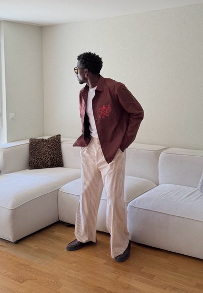 Hombre que lleva una chaqueta marrón, camisa blanca y pantalones beige a rayas, de pie frente a un sofá blanco en esquina con un cojín de estampado de leopardo.