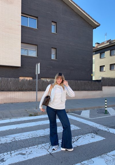 Mujer con camisa de rayas y jeans posa sobre un paso de peatones agrietado cerca de edificios residenciales bajo un cielo despejado.