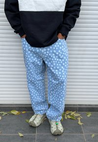 Personne portant un jean bleu clair ample à pois blancs, un sweat noir et blanc, et des baskets beiges, debout sur un sol carrelé avec des feuilles tombées.