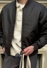 Mann in einer dunkelbraunen Lederjacke, mit einem cremefarbenen Strickpolo, grauen Hosen, der schwarze Sonnenbrille und eine cremefarbene Tragetasche hält, mit einer Uhr am Handgelenk.