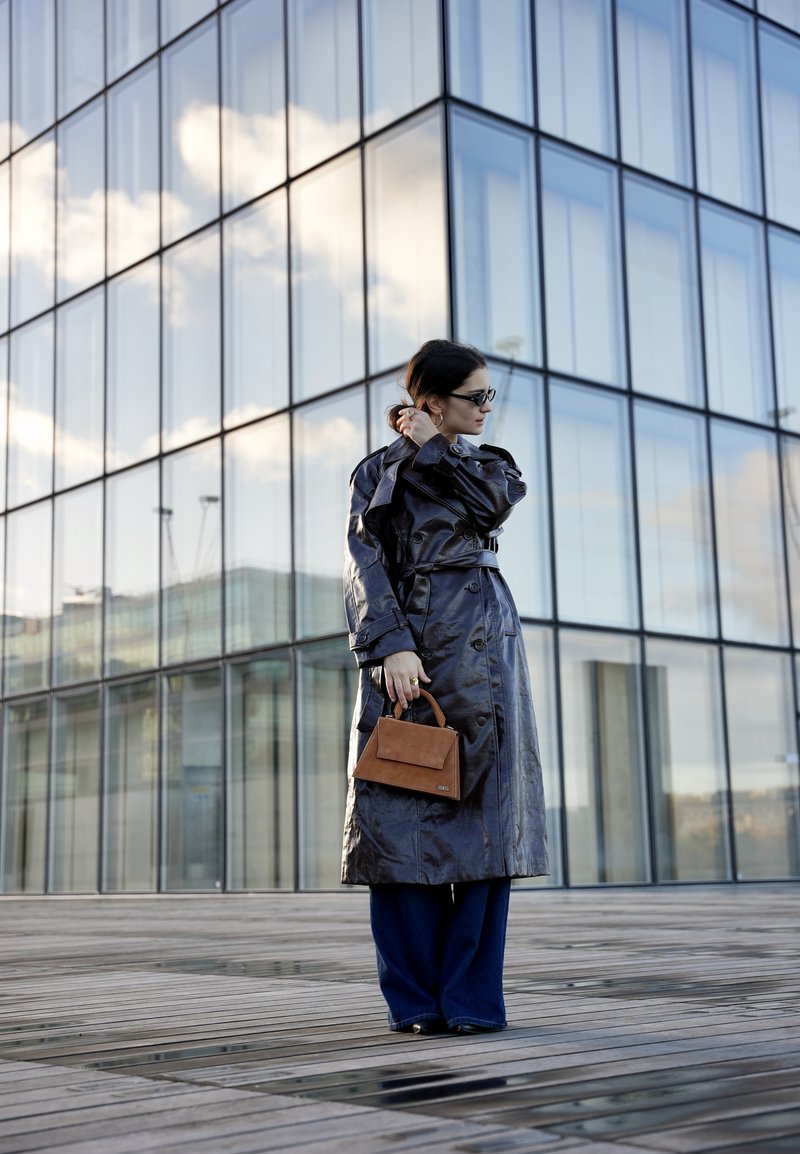 Longue trench-coat bleu foncé brillant avec une ceinture à la taille et des détails à boutons, associé à un pantalon large foncé et un petit sac à main marron.