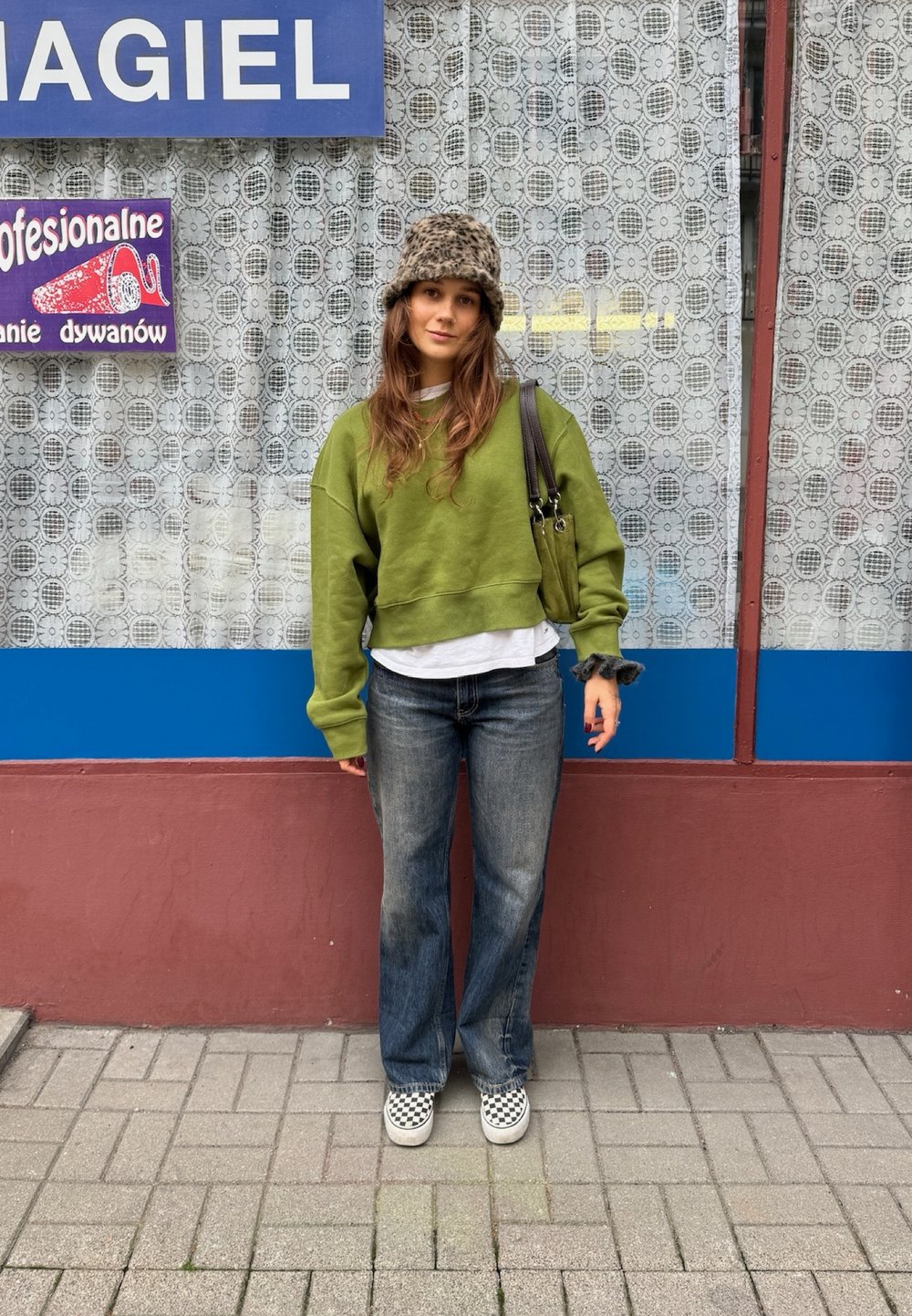 Sweatshirt verde encurtado, t-shirt branca por baixo, jeans azuis, sapatos slip-on com padrão de piso de xadrez e um chapéu com padrão de leopardo.