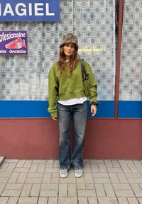 Sweatshirt verde encurtado, t-shirt branca por baixo, jeans azuis, sapatos slip-on com padrão de piso de xadrez e um chapéu com padrão de leopardo.
