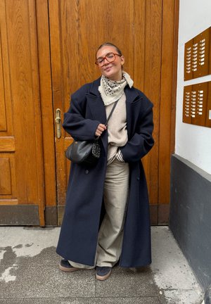 Mujer con un abrigo oscuro grande, pantalones beige, bufanda estampada y gafas, de pie con un bolso negro frente a una puerta de madera con timbres.