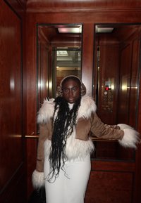 Veste en fausse fourrure et daim avec accents en fourrure blanche, associée à une jupe blanche, dans un ascenseur aux panneaux en bois. Cheveux longs et tressés avec cache-oreilles.
