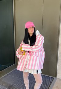 Chaqueta a rayas rosa y blanca de gran tamaño, vestido blanco, medias transparentes con estampado, gorra rosa con logo, sosteniendo un bolso dorado, de pie contra una puerta.