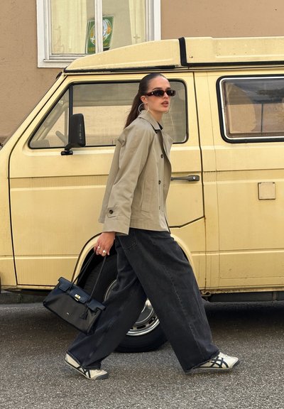 Trench beige, vaqueros de pierna ancha negros de talle alto, zapatillas grises y un bolso negro. Gafas de sol y pendientes de aro como accesorios.