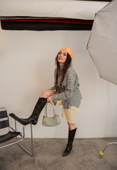 Mujer con camisa de cuadros, falda beige, botas negras hasta la rodilla y gorro naranja, sosteniendo un bolso verde y posando con un pie sobre una silla.