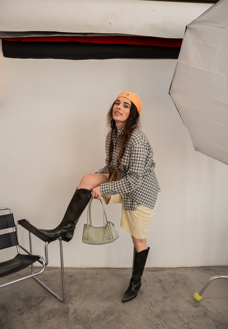Femme portant une chemise à carreaux, une jupe beige, des bottes noires montantes et un bonnet orange, tenant un sac à main vert et posant avec un pied sur une chaise.