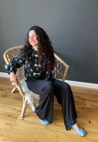 Schwarze Bluse mit floralem Muster und blauer Stickerei, graue weit geschnittene Hose und hellblaue Schuhe. Sitzt auf einem Korbsessel auf einem Holzboden.