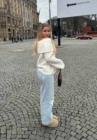 Cremfarbener schulterfreier Pullover, hellblaue weit geschnittene Jeans, sandfarbene Wildleder-Plateauschuhe, trägt eine kleine dunkle Handtasche; Kopfsteinpflaster-Hintergrund.