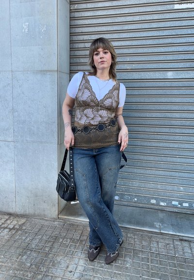 Top de encaje marrón sobre camiseta blanca, cinturón circular negro, jeans de pierna ancha azul, zapatos grises, sosteniendo un bolso pequeño negro, de pie contra una pared.
