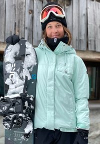 Μιντ πράσινο χιτώνιο χιονιού με κουκούλα, μαύρα γάντια, κρατάει ένα σνόουμπορντ με μαύρες βάσεις και χιόνι. Το φόντο περιλαμβάνει ξύλινες κατασκευές.