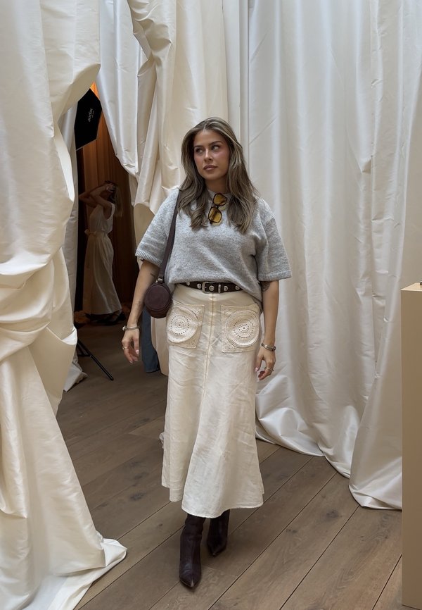 Vrouw met lang bruin haar, grijze top en crèmekleurige rok met zakken, staand op een houten vloer temidden van beige gedrapeerde gordijnen.