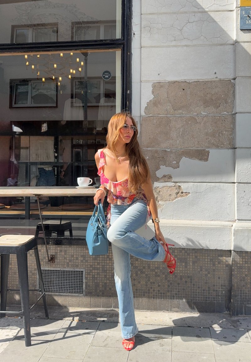 Femme en haut fleuri et jean ajustant des talons hauts rouges devant un café avec fenêtre, table, tabourets et tasse sur la table.