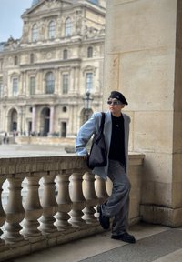 Hele helesinine bleiser, must särk, hallad teksad ja mustad kingad. Must beret ja prillid lisavad eristuvaid aksessuaare. Taustal on kivist arhitektuur.