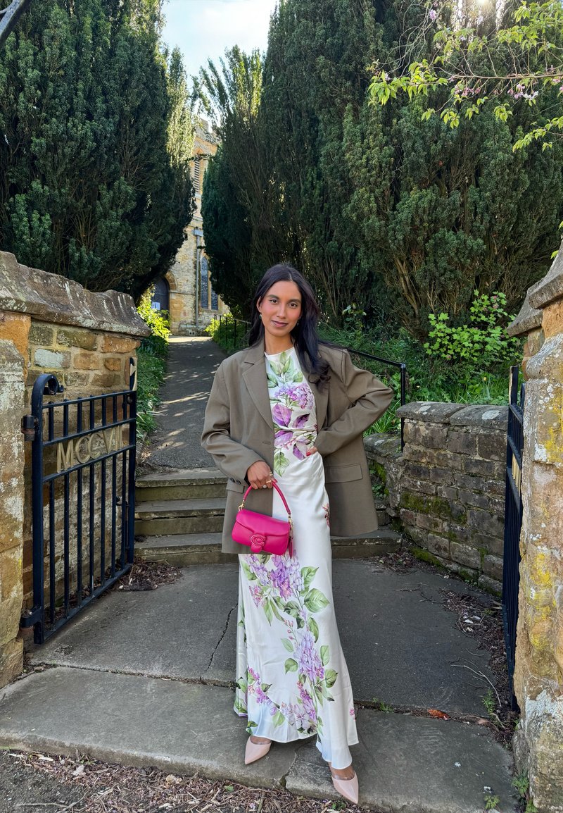 Femme en robe fleurie et blazer marron oversize tient un sac rose, debout sur un chemin entre des murs en pierre et de grands arbres près d'un passage fermé par une grille.