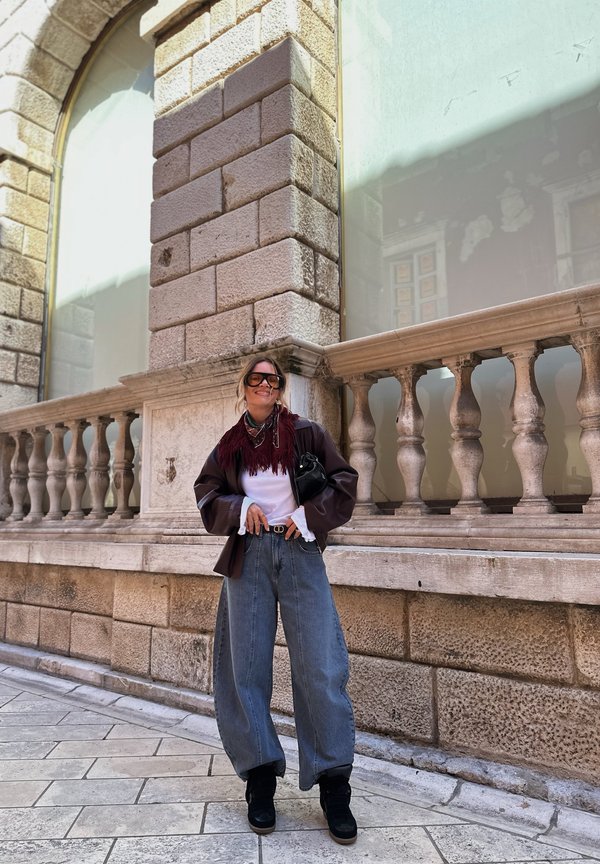 Vrouw die een oversized spijkerbroek, zwarte laarzen, een bruine jas, wit overhemd en sjaal draagt, staand tegen een stenen balustrade op een kasseienstraat.