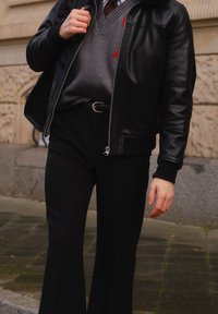 Czarna skórzana kurtka bomberka, szary sweter z dekoltem w serek z czerwonymi detalami, czarne rozkloszowane spodnie oraz srebrna sprzączka paska. Teksturowane materiały, casualowy design.