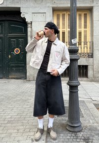 Krēmveida žakete ar rozā logo, melna T-kreklu, melni džinsu kulotu, bēšas kurpes un baltas zeķes; tur rokās kafijas krūzi ārpus telpām pie pelēkām durvīm.