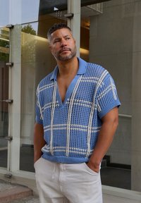 Zila adīta polo krekls ar V veida kaklu, kuram ir krēmkrāsas režģa raksts. Valkāts ar baltām biksēm, kas izceļ teksturētu, atvērtu dizainu.