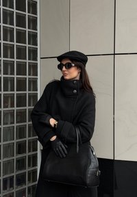 Svarte ullfrakk med høy krage og knapper, svart flat caps, store solbriller, svarte skinnhansker og en teksturert svart veske.