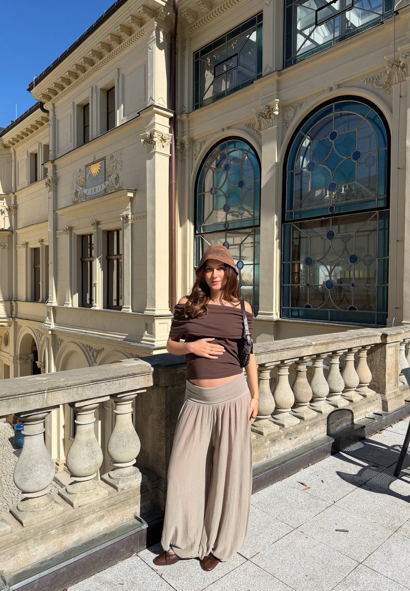 Femme en haut marron et pantalon large beige se tenant sur un balcon en pierre ensoleillé avec une balustrade ornée et de grandes fenêtres en arc derrière.