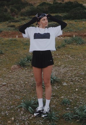 Joven mujer al aire libre ajustándose una gorra negra, vistiendo una camiseta blanca corta, pantalones cortos negros, mangas largas negras en los brazos, calcetines blancos y zapatillas negras.
