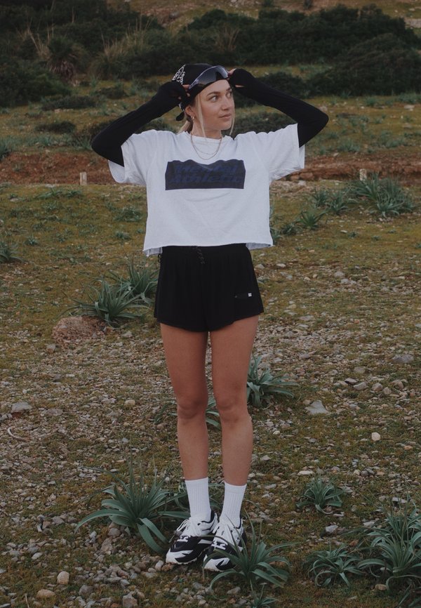 Mlada ženska na prostem, ki si prilagaja črno kapo, nosi bel odrezan T-shirt, črne kratke hlače, dolge črne rokavice, bele nogavice in črne superge.