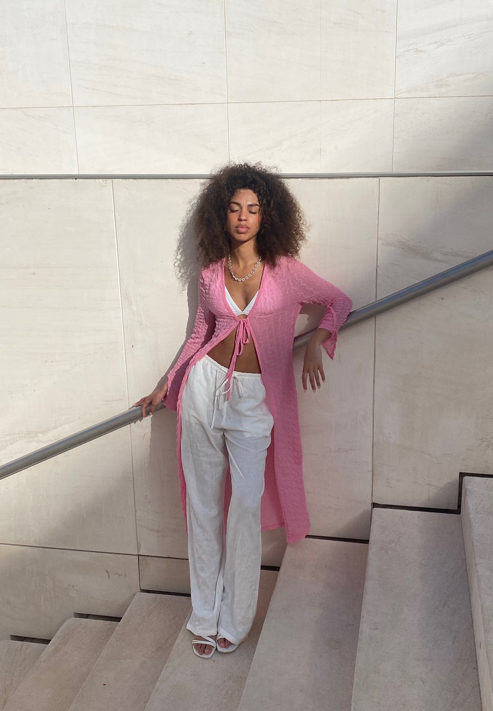 Lång rosa kofta med texturerad tyg, knuten i midjan, bärs över en vit topp och lösa vita byxor, parat med vita sandaler.