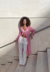 Lång rosa kofta med texturerad tyg, knuten i midjan, bärs över en vit topp och lösa vita byxor, parat med vita sandaler.