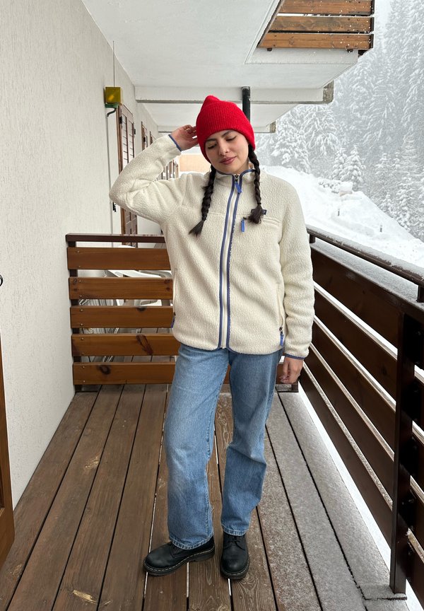 Femme aux cheveux tressés portant un bonnet rouge, une veste en polaire blanche, un jean et des bottes noires, debout sur un balcon en bois avec des arbres enneigés en arrière-plan.