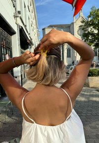 Persona con capelli castano chiaro, lunghi fino alle spalle, legati indietro con una clip gialla, che indossa un top bianco senza maniche, in piedi all'aperto.