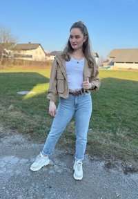 Casaco cropped bege com bolsos sobre uma camisa branca canelada, combinado com jeans claros de cintura alta e sapatilhas brancas chunky.