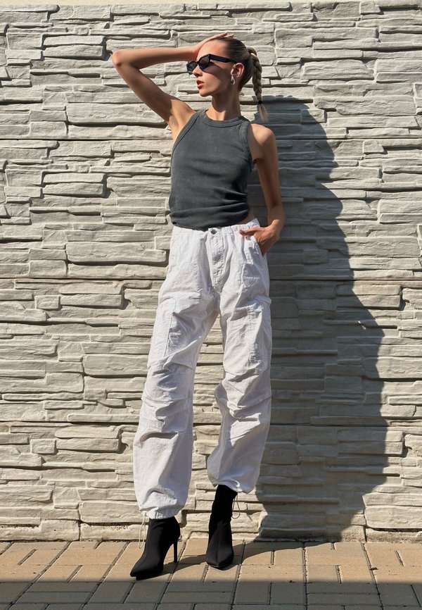 Grå ærmeløs tanktop, hvide cargo-bukser med lommer og sorte ankelstøvler. Tekstureret, struktureret vægbaggrund.