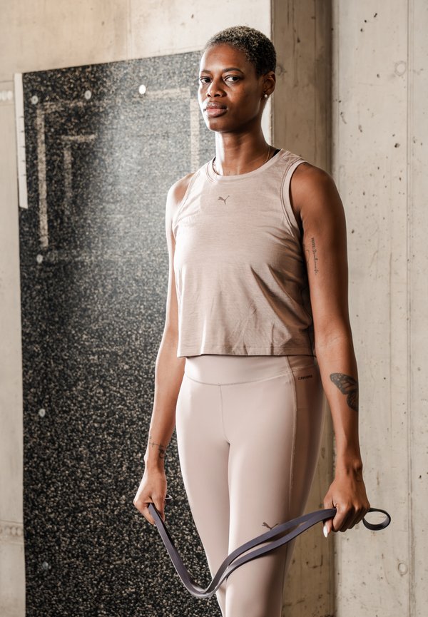 Femme athlétique en tenue de sport beige tenant une bande de résistance, debout dans une salle de sport avec des murs en béton et du matériel d'entraînement en arrière-plan.