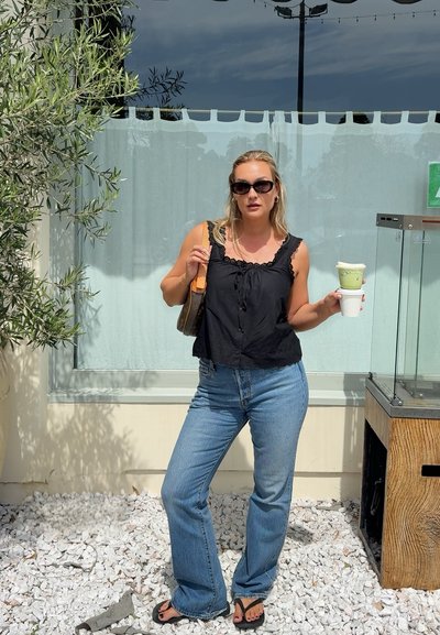 Mujer con top negro sin mangas, vaqueros azules, gafas de sol, sostiene dos bebidas, de pie sobre grava blanca junto a un edificio con plantas cercanas.