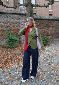 Grønn blazer med tynne striper, rød strikket skjerf, vide mørke denimbukser og hvite sko. Bærer en lys rosa håndveske. Brune blader spredt på bakken.