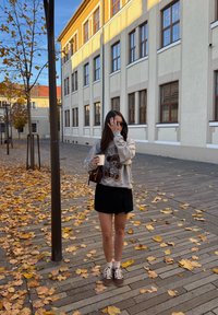 Hanorac gri cu grafică de ursuleț de pluș, fustă neagră și adidași maro. Ținând o cană de cafea, stând printre frunze galbene căzute pe trotuar.