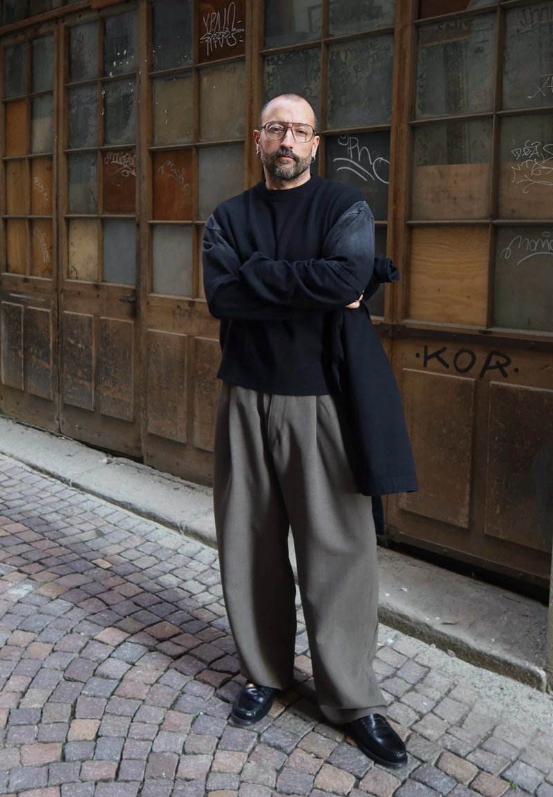 Homme avec lunettes, barbe et tête rasée, debout les bras croisés, portant un pull noir et un pantalon large gris dans une rue pavée.