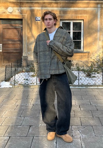 Chaqueta de cuadros, de gran tamaño, en verde apagado y marrón, llevada sobre una camisa blanca. Jeans negros holgados, zapatos beige. Llevando un bolso de bandolera verde.