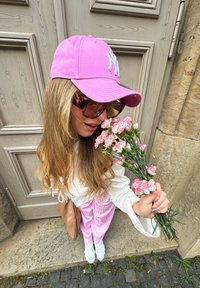 Gorra rosa, gafas de sol oversized, camiseta blanca de manga larga, pantalones a rayas rosas, zapatillas blancas, sosteniendo flores rosas, frente a una puerta de madera.