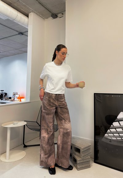 Camiseta blanca corta y pantalones amplios marrón texturizados; sosteniendo una taza; entorno minimalista con acentos geométricos en negro y gris.