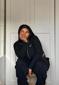 Μαύρη φούτερ ζακέτα με λογότυπο Nike, σε συνδυασμό με χαλαρές μαύρες παντελόνες. Το υποκείμενο κάθεται σε μια ξύλινη σκάφη, με πλεξούδες και παιχνιδιάρικη έκφραση.