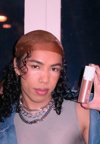 Een persoon met krullend zwart haar in een bruine hoofddoek houdt een cilindervormige fles met een witte dop vast, terwijl hij zilveren kettingen en een grijs topje draagt.