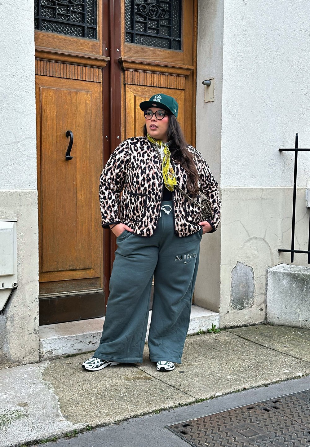 Leopardenmusterjacke mit schwarzem Rand, grüne übergroße Hose mit Logo, grüne Mütze und Turnschuhe mit kontrastierenden Schwarz-Weiß-Mustern.