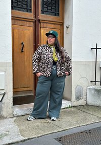 Leopardenmusterjacke mit schwarzem Rand, grüne übergroße Hose mit Logo, grüne Mütze und Turnschuhe mit kontrastierenden Schwarz-Weiß-Mustern.