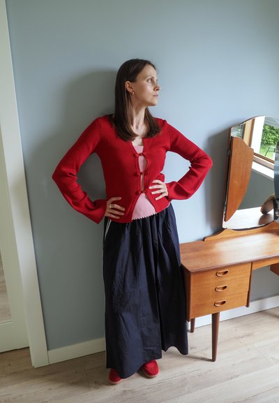 Cárdigan rojo de canalé con botones, top rosa y falda larga negra, combinados con zapatos rojos. Mesa lateral de madera de estilo mid-century en el fondo.