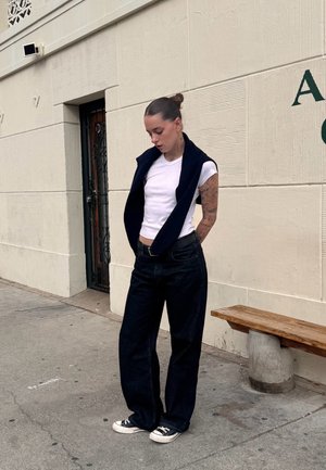 Una camiseta blanca ajustada debajo de una chaqueta sin mangas azul marino, combinada con unos jeans de pierna ancha oscuros y zapatillas Converse negras. El fondo presenta una pared texturizada.