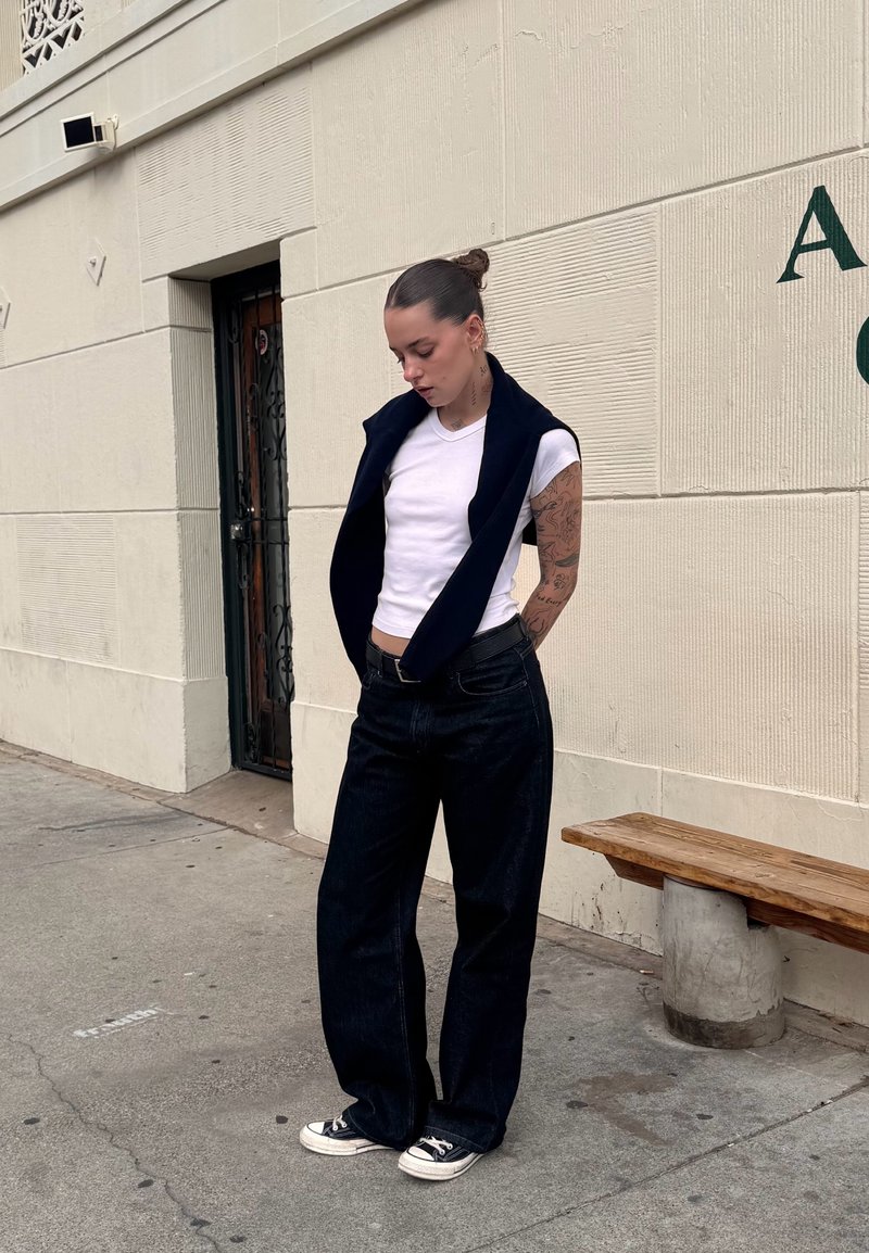 White fitted t-shirt worn under a navy sleeveless jacket, teamed with dark wide-leg jeans and black Converse sneakers. The background showcases a textured wall.